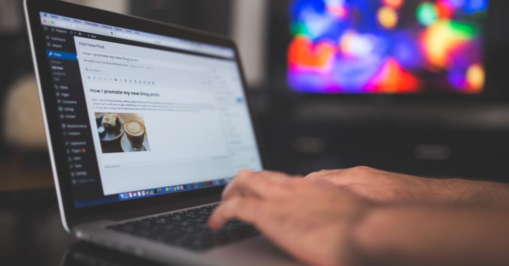Hands typing on a laptop with a blog post visible, cozy indoor setting with colorful screen in background.